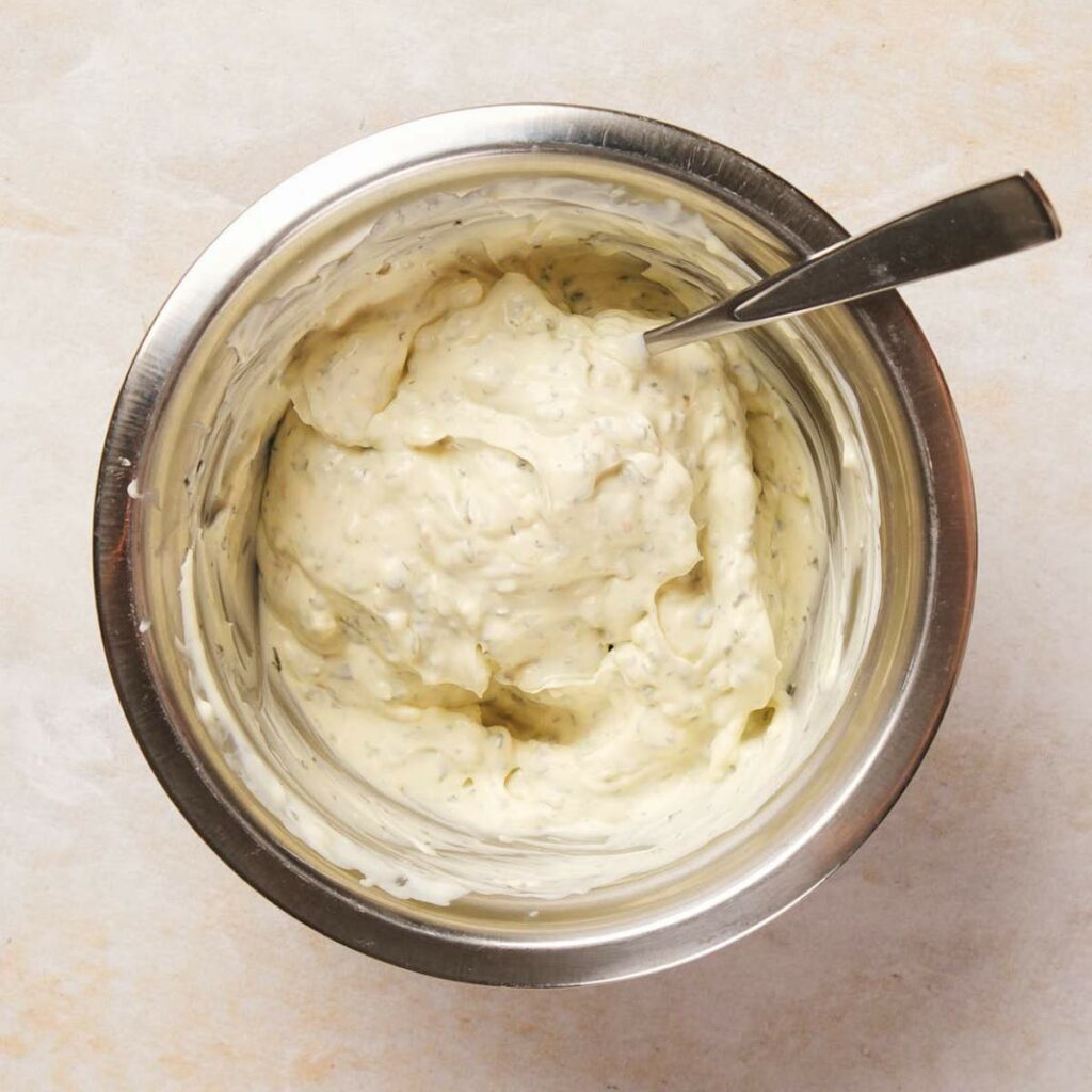 Bowl filled with creamy mayonnaise-based sauce with herbs, with a spoon resting inside.