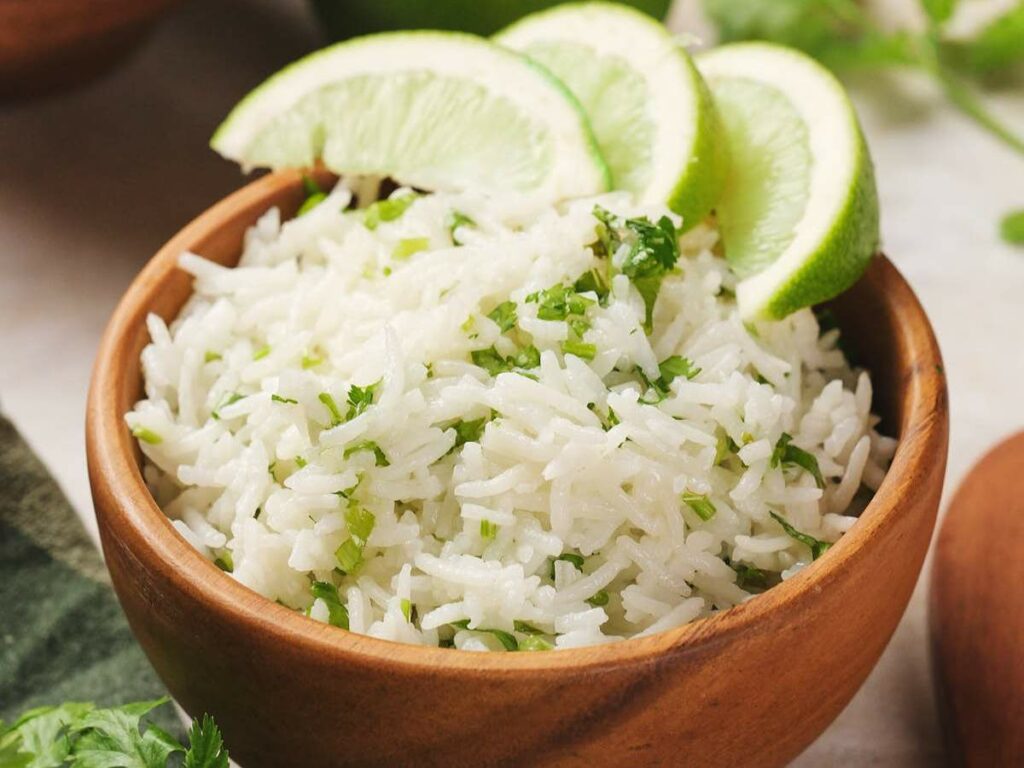 Wooden bowl filled with white rice garnished with chopped cilantro and three lime wedges on top.