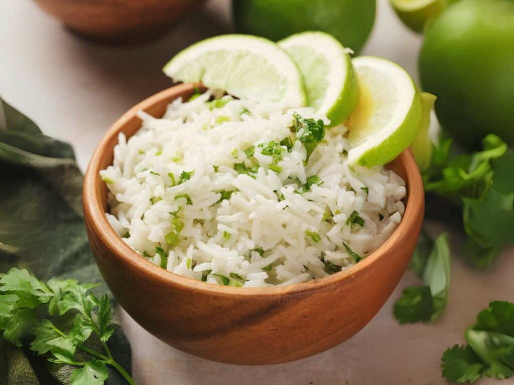 A wooden bowl of white rice garnished with fresh cilantro and lime slices, surrounded by cilantro leaves and whole limes.