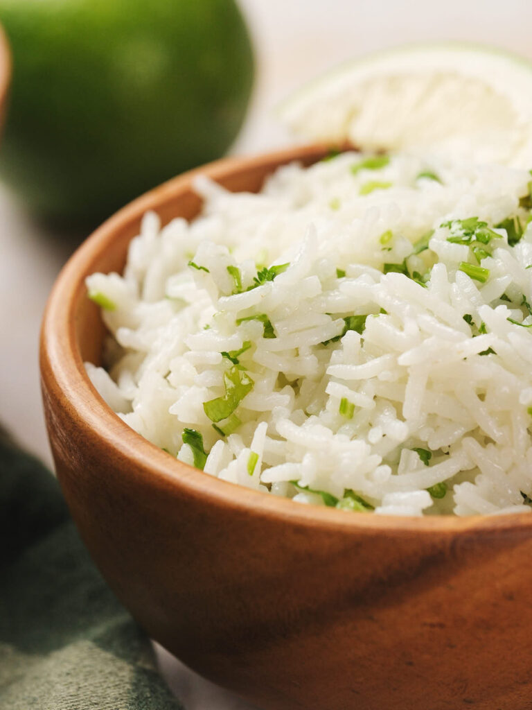 A wooden bowl filled with cilantro lime rice garnished with chopped herbs, with a lime wedge in the background.