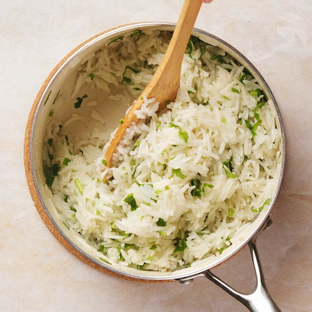 Cooked rice in a pot being stirred with a wooden spoon, with visible green herbs mixed in.