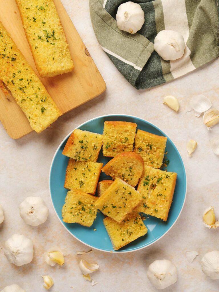 Slices of garlic bread on a blue plate surrounded by whole garlic bulbs and cloves.