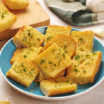 A plate of sliced garlic bread topped with parsley. Garlic bulbs and slices are nearby, with a cloth and wooden board in the background.