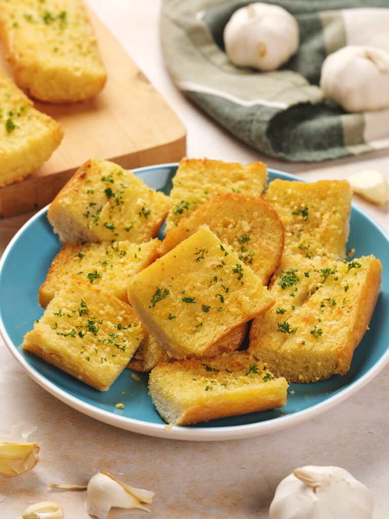 A plate of sliced garlic bread topped with parsley. Garlic bulbs and slices are nearby, with a cloth and wooden board in the background.