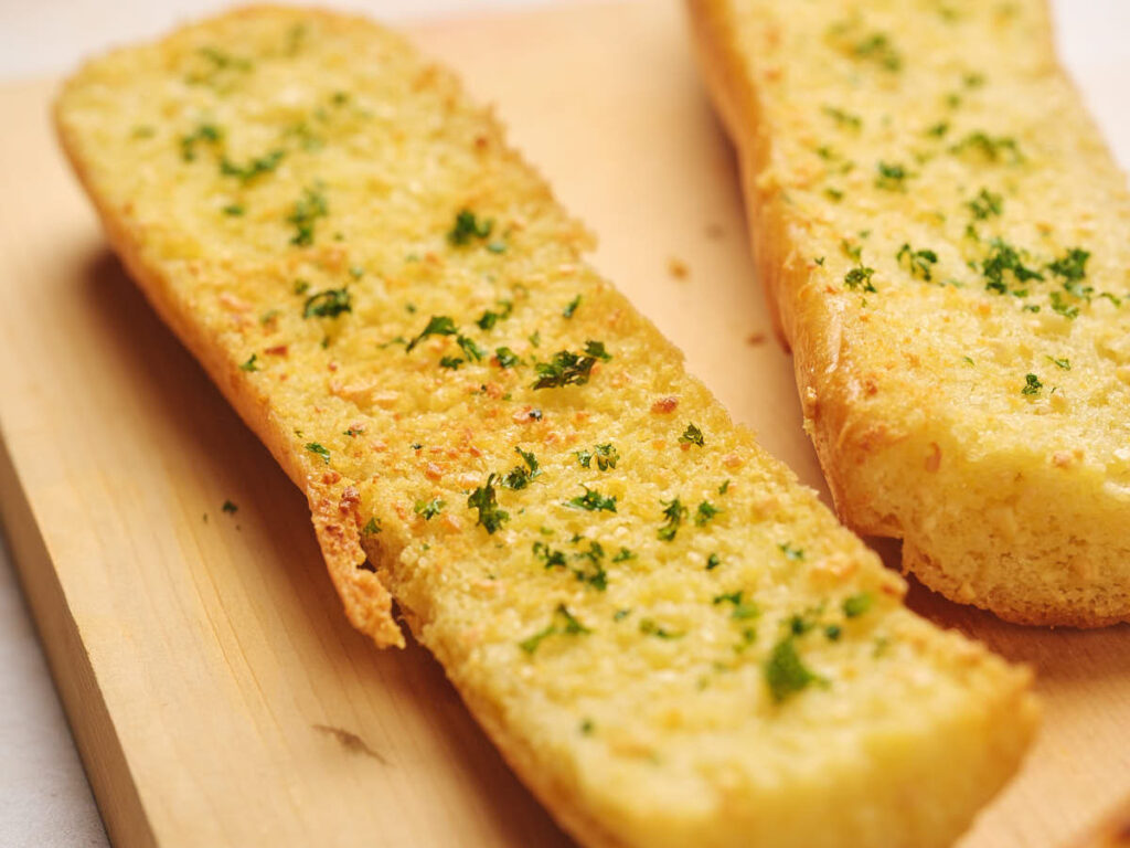 Two slices of garlic bread topped with chopped parsley on a wooden surface.