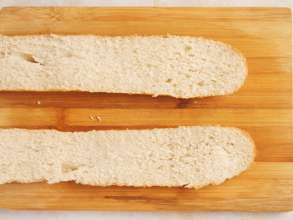 Two halves of a sliced baguette are placed side by side on a wooden cutting board.