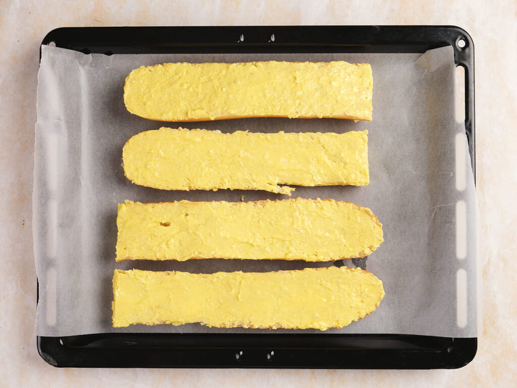 Four long slices of bread coated with yellow spread on a baking tray lined with parchment paper.