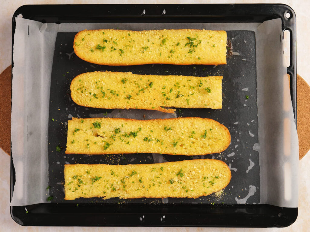 Four pieces of garlic bread with parsley on a parchment-lined baking tray.