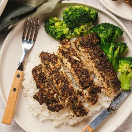 Sliced herbed chicken breast on white rice with broccoli on a plate, accompanied by a fork and knife.