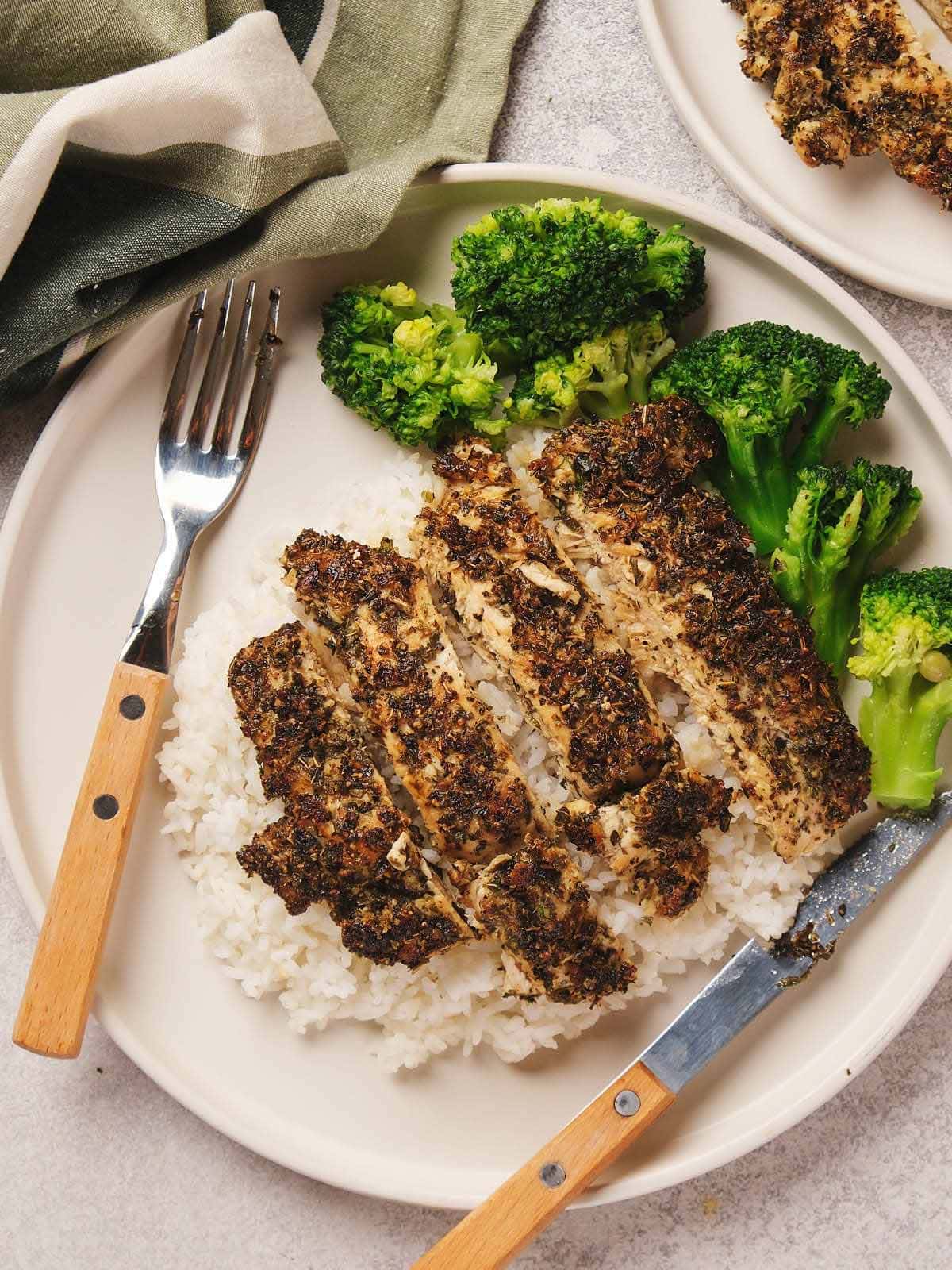 Sliced herbed chicken breast on white rice with broccoli on a plate, accompanied by a fork and knife.