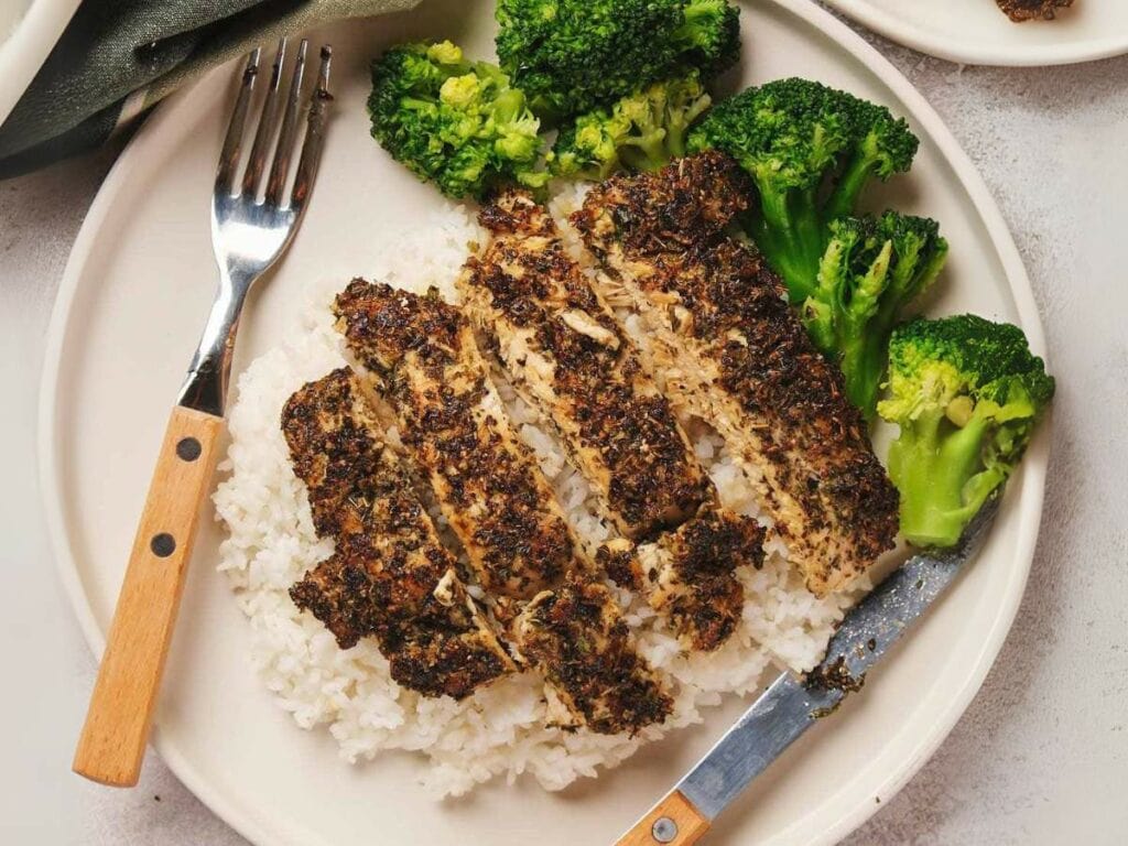 Herb-crusted sliced chicken served on white rice with steamed broccoli, accompanied by a fork and knife on a white plate.