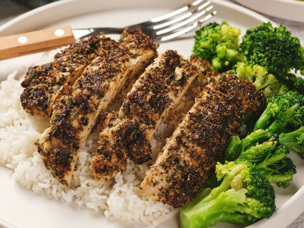 Sliced herb-crusted chicken on rice with broccoli on a white plate, accompanied by a fork.