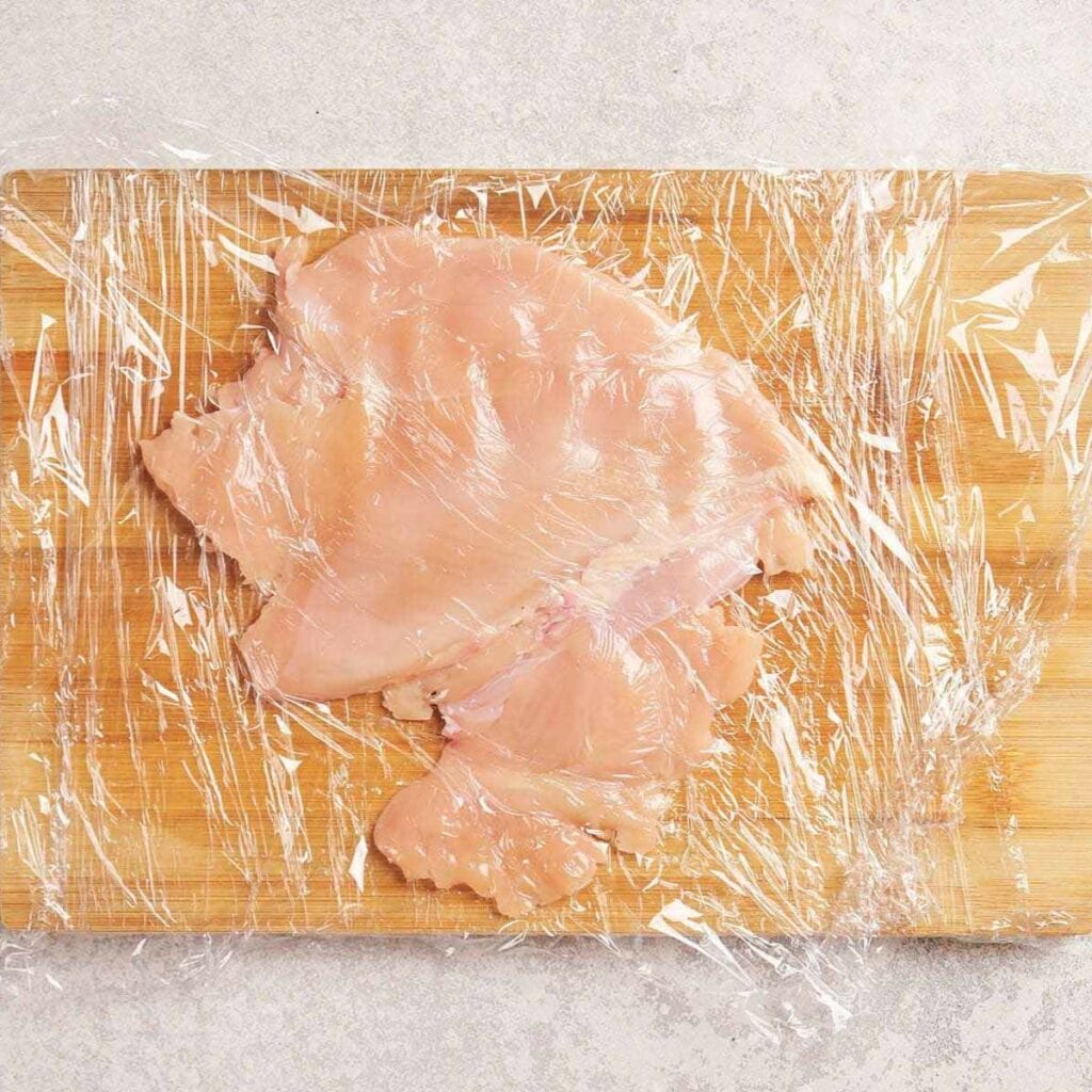 Flattened raw chicken breast covered with plastic wrap on a wooden cutting board.