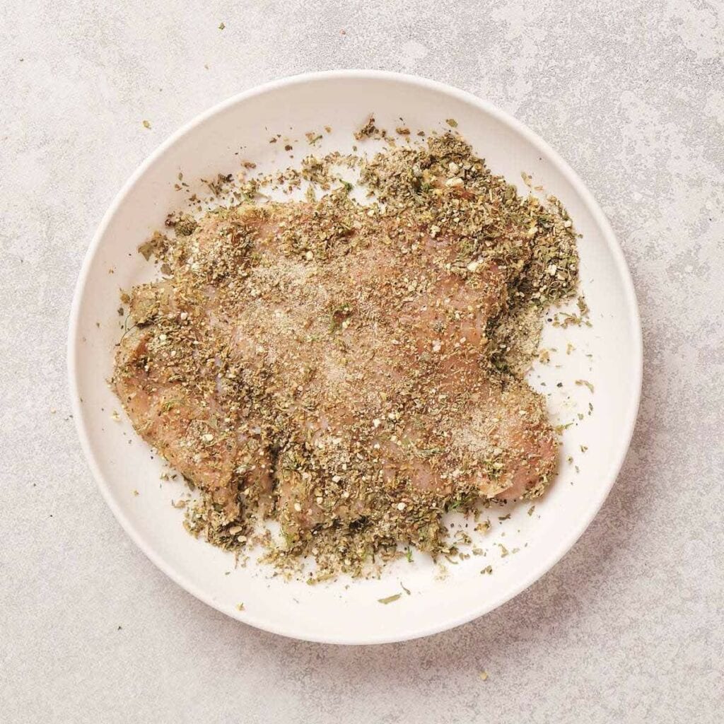 Raw chicken breast coated with a mixture of herbs and spices on a white plate.