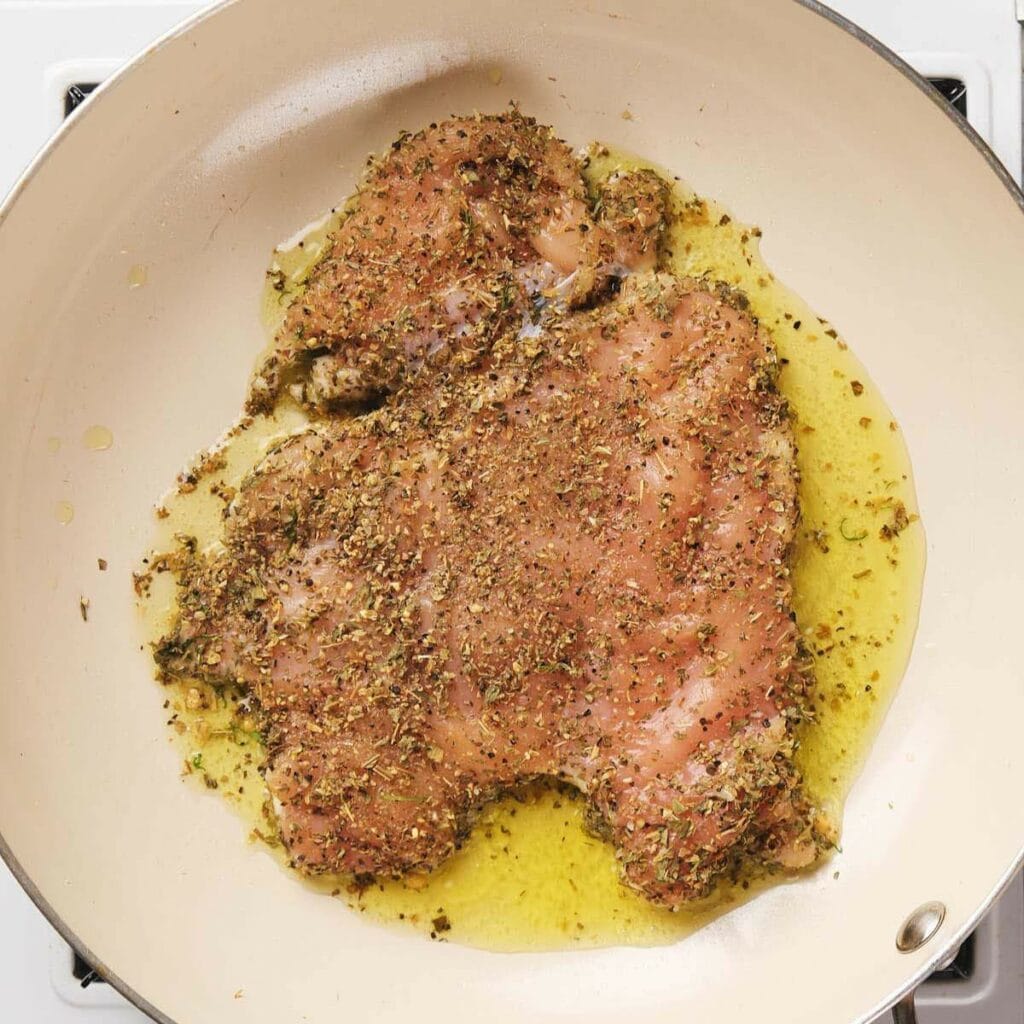 Seasoned chicken breast cooking in a pan with oil.