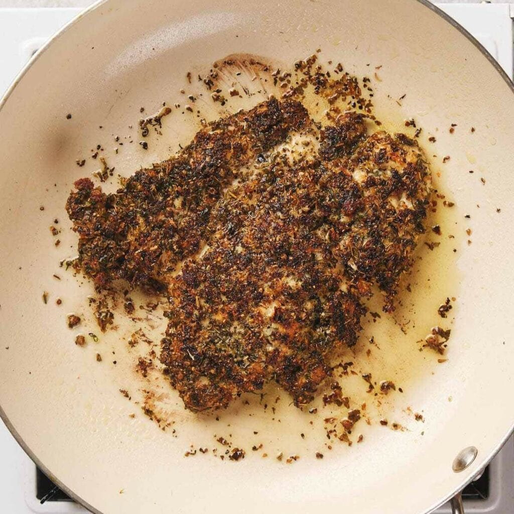 Chicken breast coated with herbs and spices, cooking in a beige frying pan.