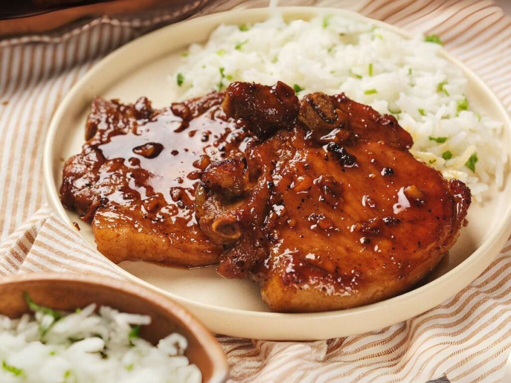 Two honey glazed pork chops on a plate next to a serving of white rice with herbs, placed on a striped cloth.