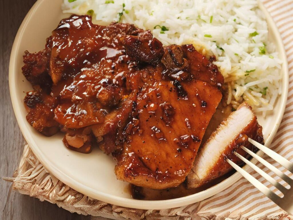 Plate with sliced honey glazed pork chops and a side of white rice garnished with herbs.