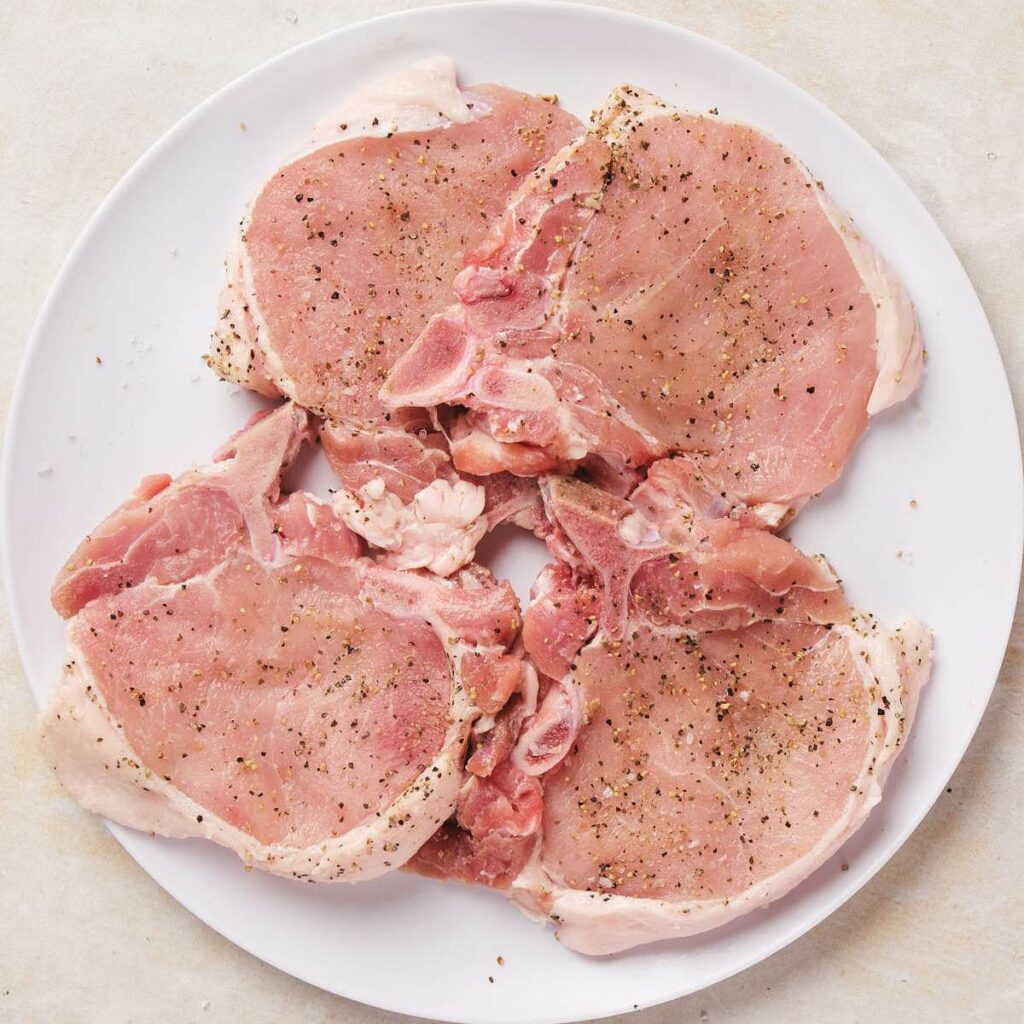 Four raw pork chops seasoned with pepper on a round white plate.