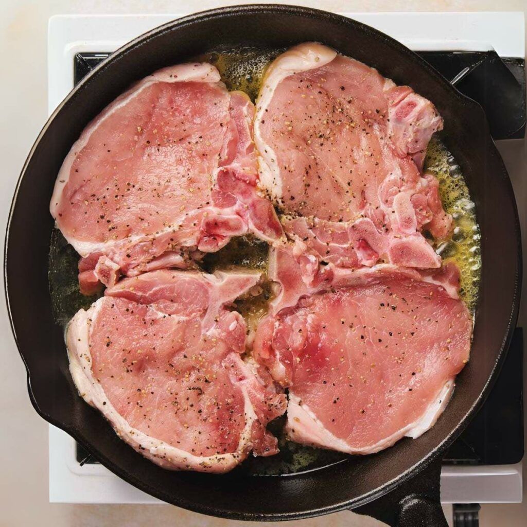 Four raw pork chops seasoned with black pepper are sizzling in a black skillet on a stovetop.