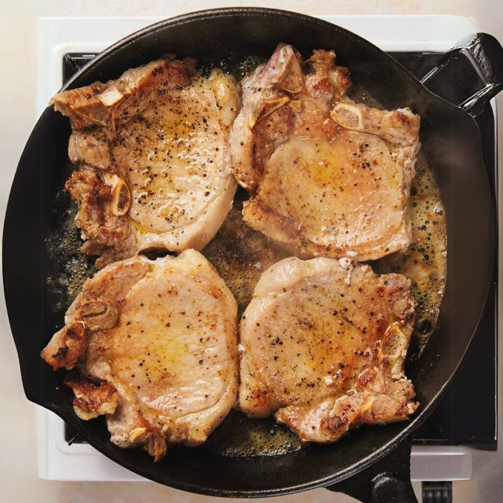 Four seasoned pork chops being pan-fried in a black skillet on a stovetop.