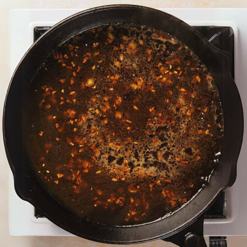 A cast iron skillet on a stove is filled with a bubbling brown liquid containing visible pieces of garlic or similar ingredients.