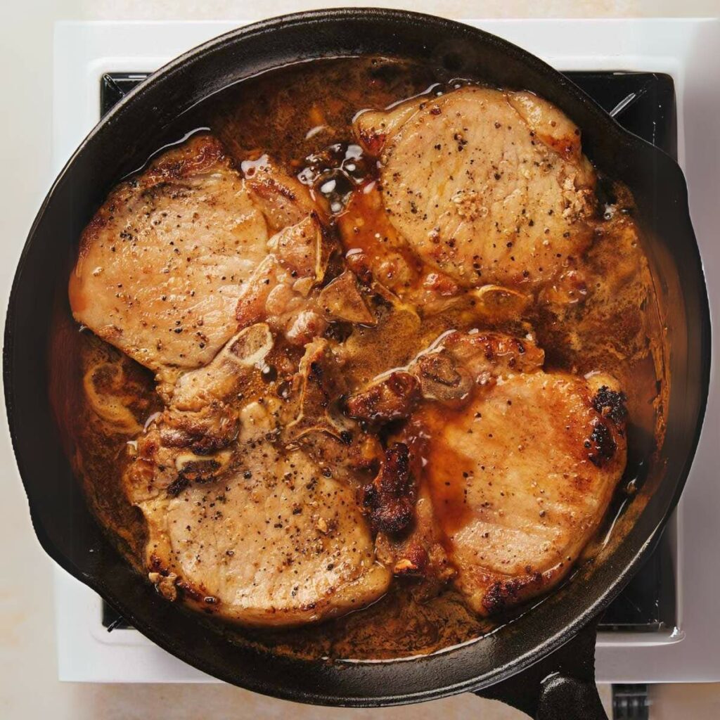 Pork chops cooking in a black skillet on a stovetop. The meat is browned and seasoned, surrounded by juices.