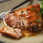 A plate with glazed, partially eaten chicken thigh garnished with herbs, accompanied by a fork and some green garnish on a light-colored dish.