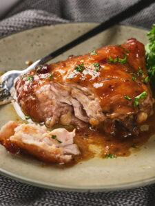 A plate with glazed, partially eaten chicken thigh garnished with herbs, accompanied by a fork and some green garnish on a light-colored dish.