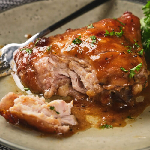 A plate with glazed, partially eaten chicken thigh garnished with herbs, accompanied by a fork and some green garnish on a light-colored dish.