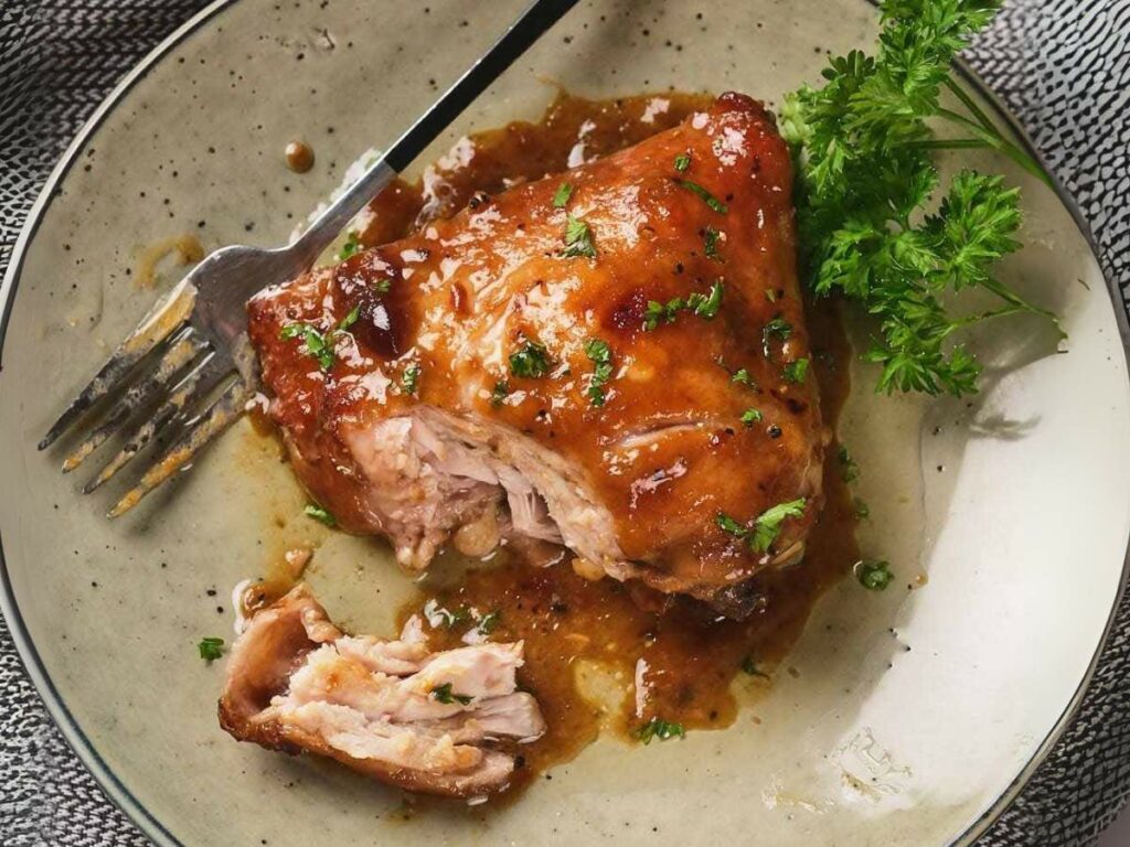 A plate with apricot chicken thigh garnished with herbs. A fork rests beside a piece of torn chicken.