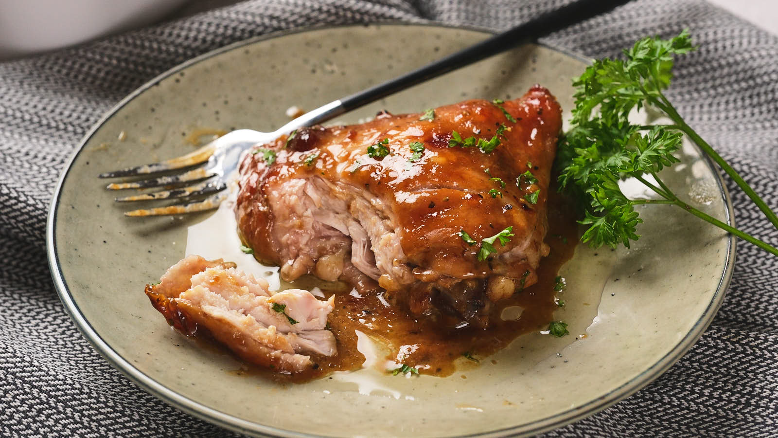 A glazed chicken thigh on a green plate, garnished with parsley, alongside a fork and a piece cut off.