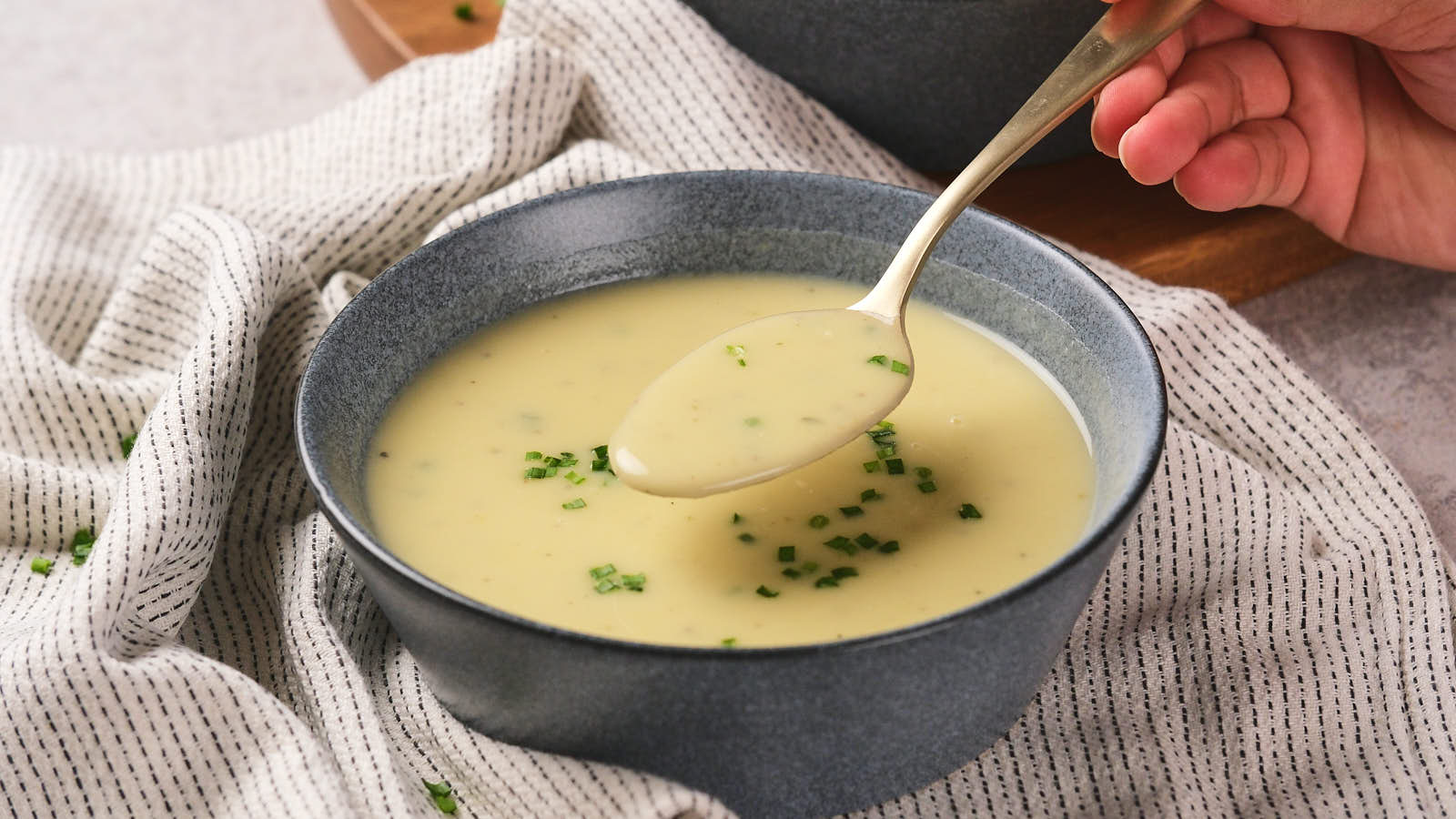 A bowl of creamy potato and leek soup garnished with chopped herbs, with a hand holding a spoon above it.