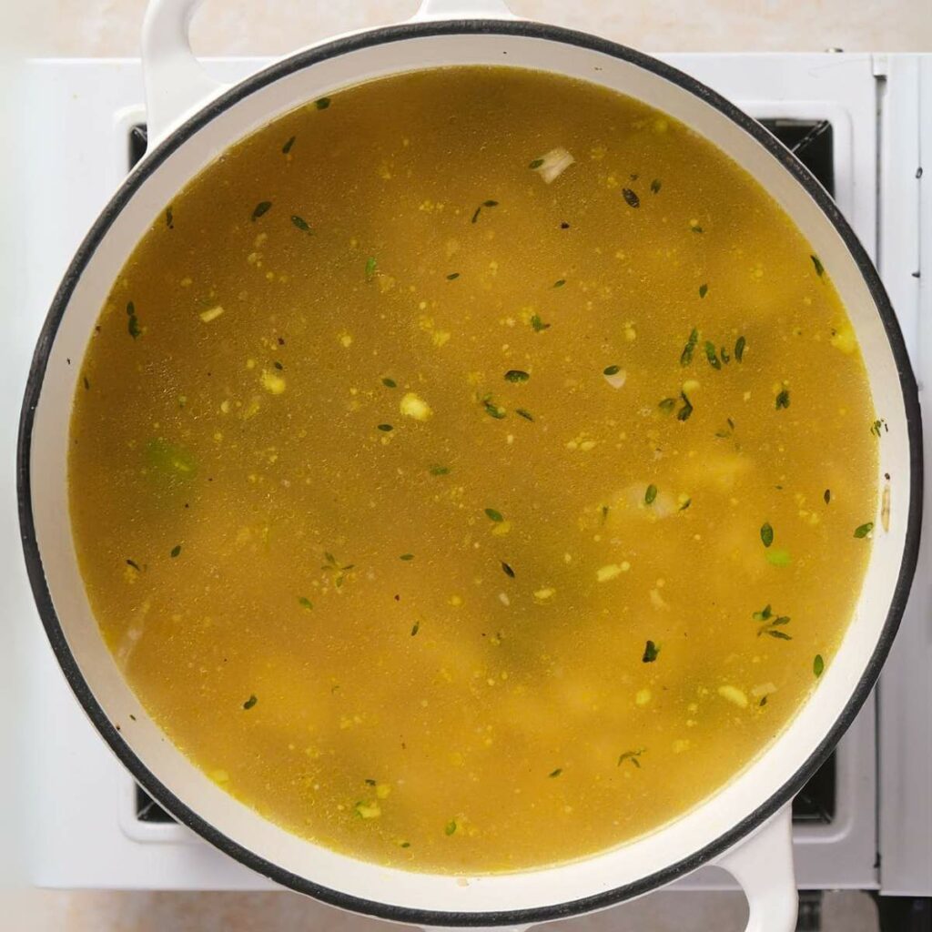 A pot of soup on the stove, filled with a yellowish broth containing herbs and small chunks of vegetables.