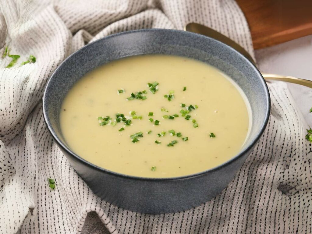 A blue bowl filled with creamy potato and leek soup garnished with chopped herbs, placed on a patterned fabric.