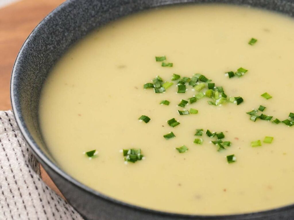 A bowl of creamy potato and leek soup garnished with chopped green herbs, placed on a striped cloth.