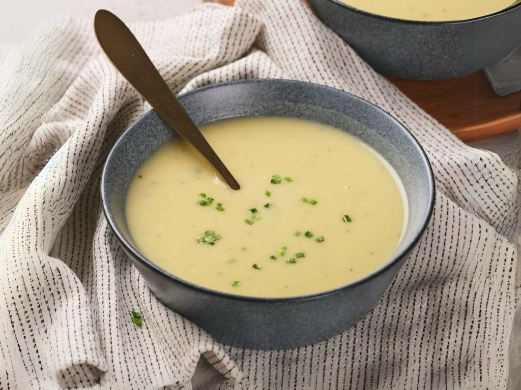 A bowl of creamy potato and leek soup garnished with chopped chives and a spoon resting inside, placed on a patterned cloth.