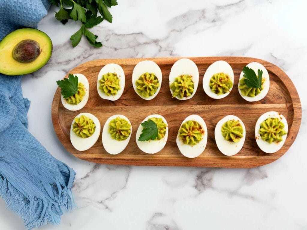 Avoacdo deviled eggs with avocado filling, garnished with parsley, arranged on a wooden platter on a marble surface.