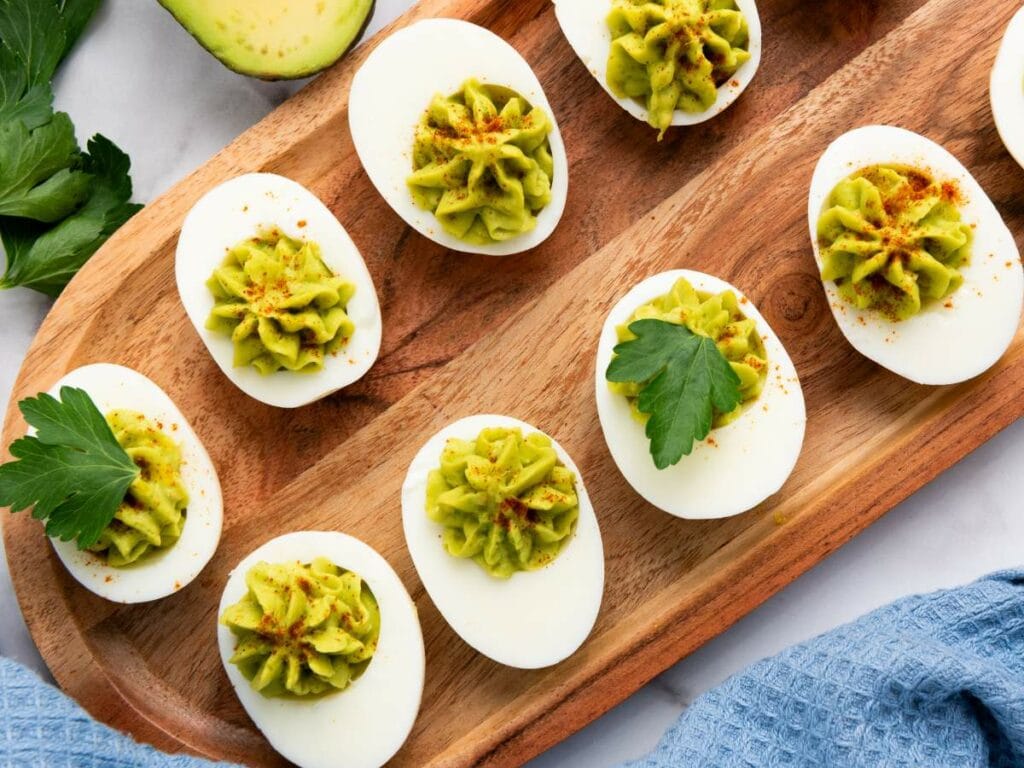 Avocado deviled eggs topped with guacamole and paprika arranged on a wooden platter, garnished with parsley.