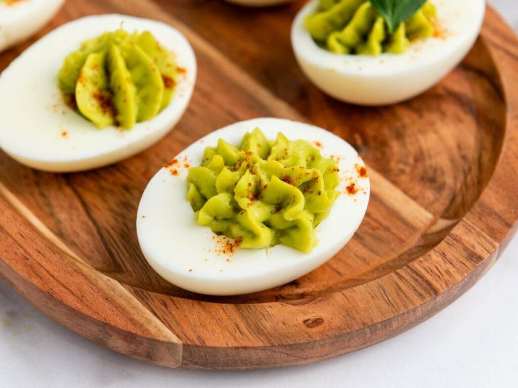 Avocado deviled eggs on a wooden plate, topped with green filling and a sprinkle of paprika.