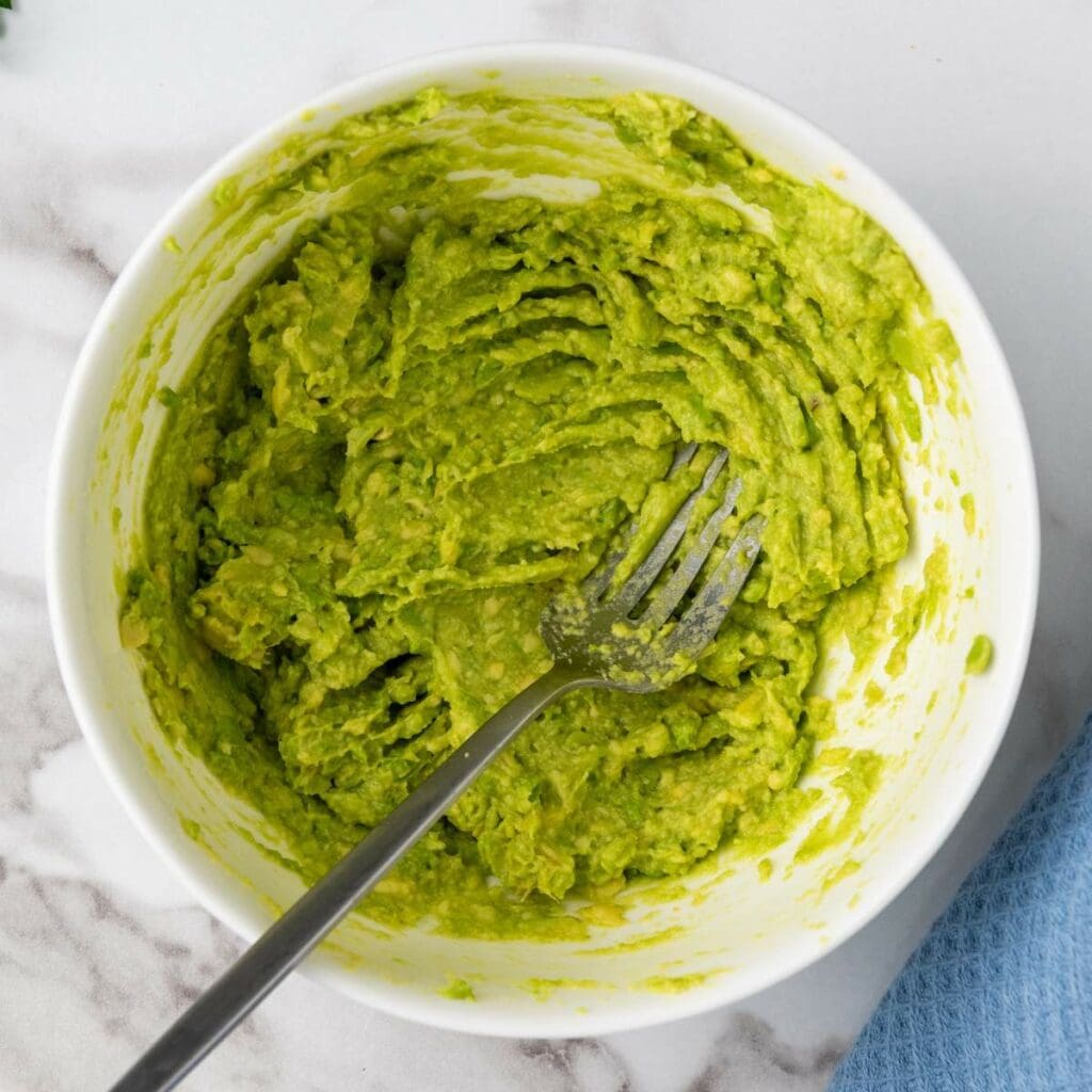 A bowl of mashed avocado with a fork inside, on a marble surface.