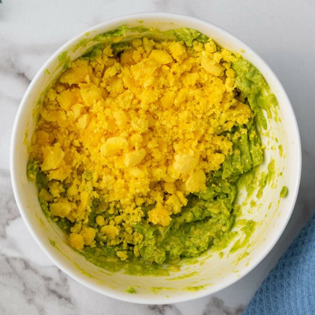 A bowl filled with mashed avocado and crumbled yellow egg yolk on a marble surface.