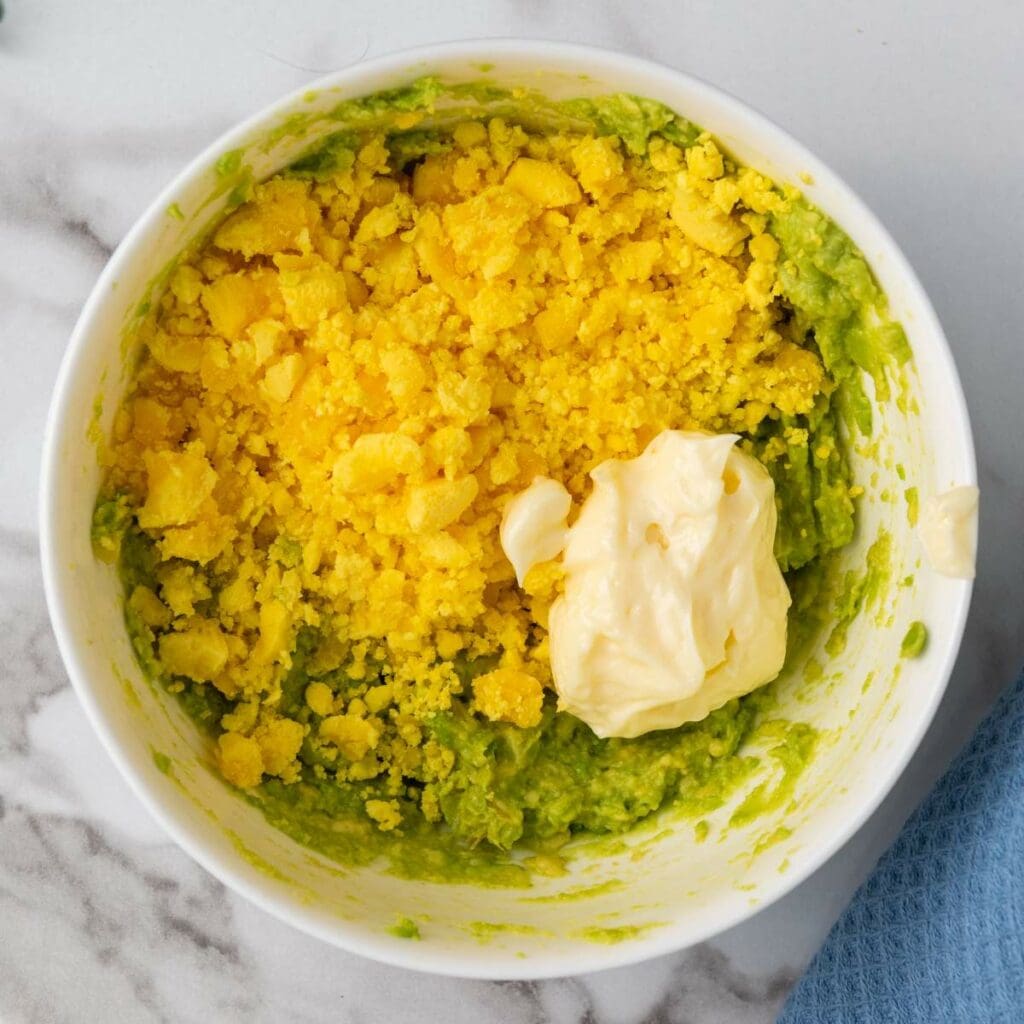 A bowl containing mashed avocado, crumbled boiled egg yolk, and a dollop of mayonnaise.