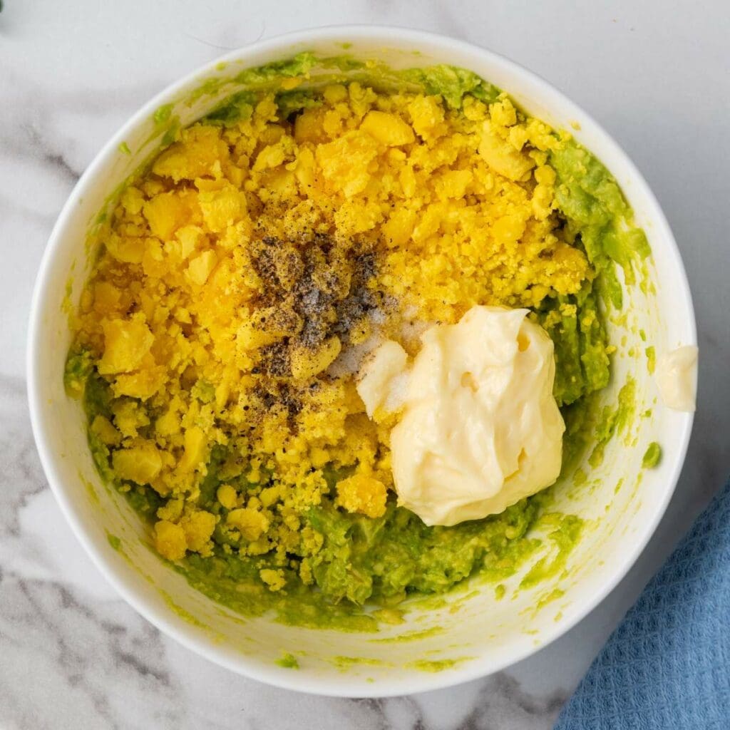 Bowl of mashed avocado mixed with crushed boiled egg, black pepper, salt, and a dollop of mayonnaise on a marble surface.