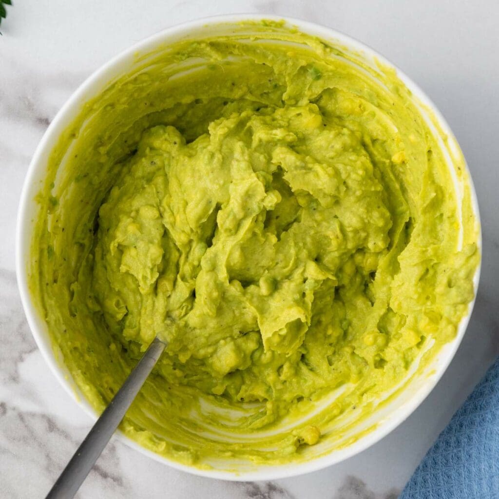 A bowl of mashed guacamole with a spoon on a marble surface.