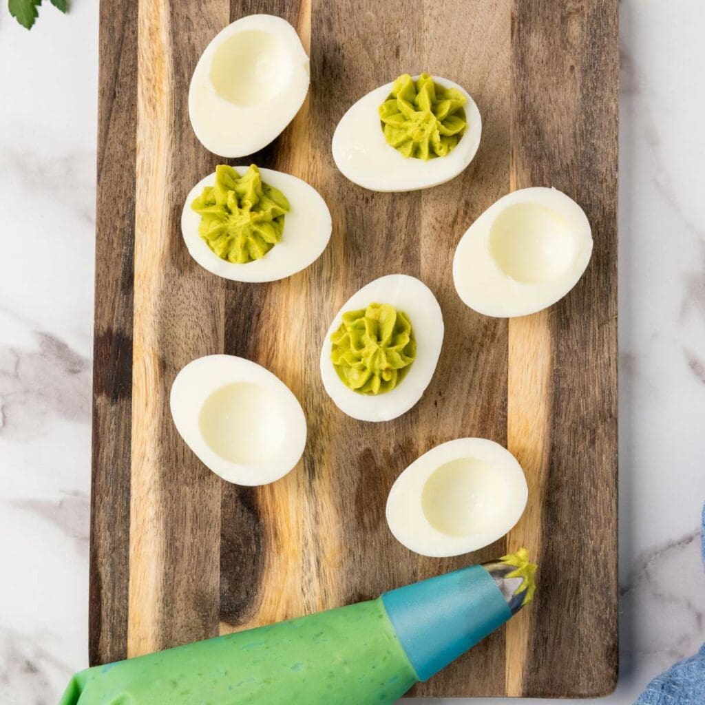 Deviled eggs being filled with a green mixture from a piping bag on a wooden cutting board.