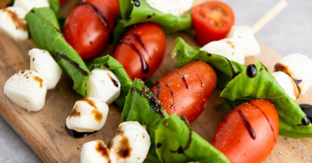 Caprese skewers with cherry tomatoes, fresh basil, mozzarella balls, and balsamic glaze on a wooden board.