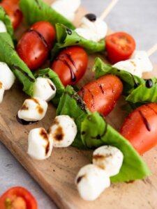 Skewers with cherry tomatoes, fresh basil, and mozzarella balls, drizzled with balsamic glaze, displayed on a wooden board.