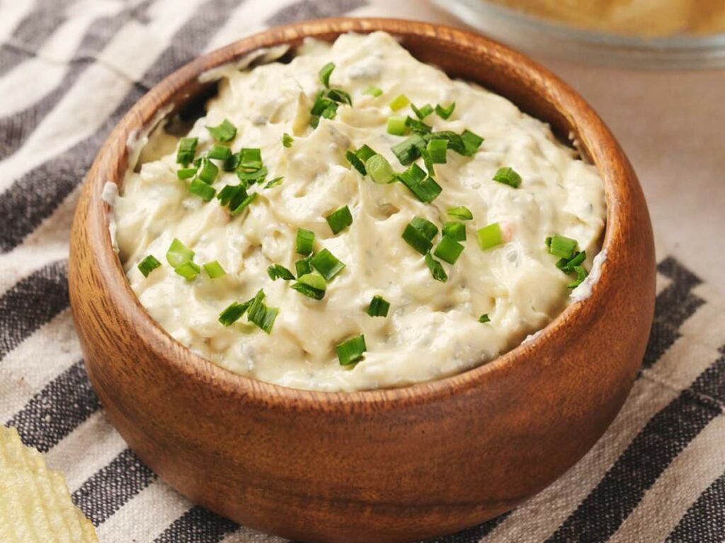 A wooden bowl filled with creamy chip dip, topped with chopped green onions, placed on a striped cloth.