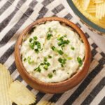 A wooden bowl of creamy white dip garnished with chopped chives, next to crinkle-cut potato chips on a striped cloth.